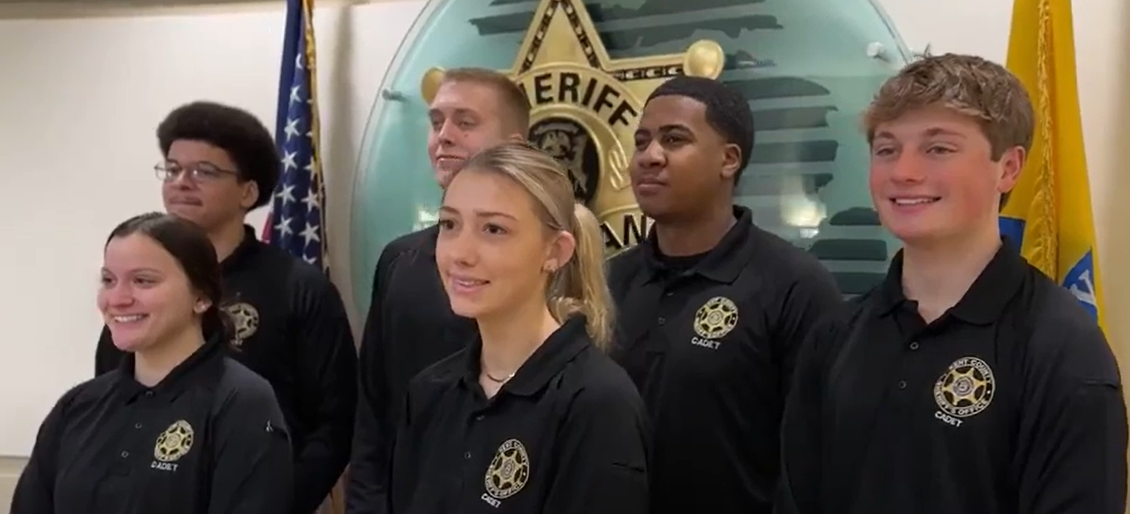 Four cadets stand in front of the Michigan Sheriff's sheild and smile at the camera